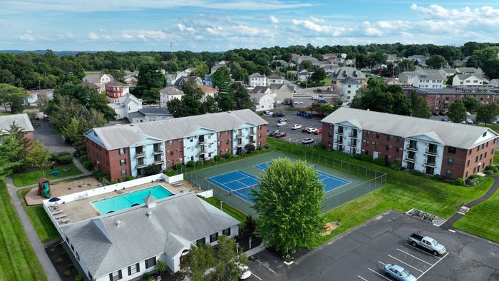 Aerial view of Princeton Place Apartments in Worcester, MA, featuring brick buildings, parked cars, a swimming pool, and tennis court, all nestled among trees and residential houses under a partly cloudy sky.