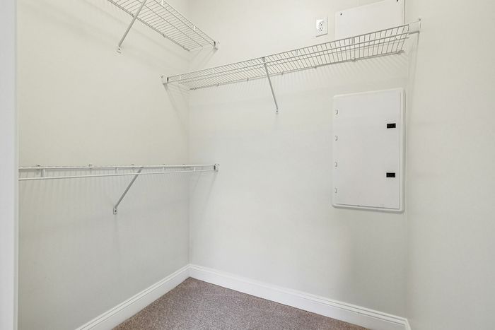 Spacious walk-in closet in an apartment in Westford, MA, featuring beige carpet, white walls, wire shelving on two sides, and an electrical panel on the right. The space is well-lit and clean. | Apartments with Walk-in closets in westford  Princeton Westford | Princeton Westford Apartments