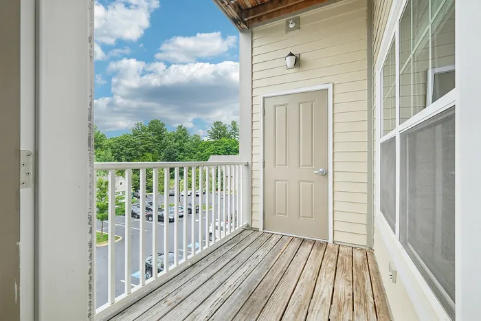 A small private balcony with wooden flooring, beige walls, and a closed door overlooks a parking lot and green trees under a partly cloudy sky. White railing lines the balcony&rsquo;s edge in this apartment in Westford, MA. | Apartments with Balcony in Westford