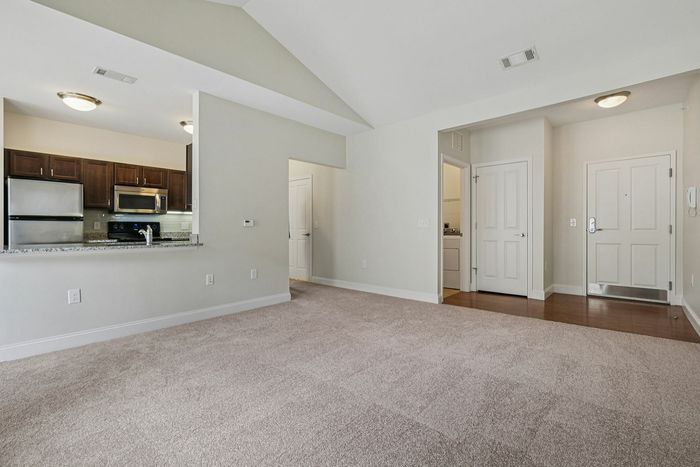 Bright, empty apartment in Westford MA featuring a spacious living room with beige carpet, adjacent to an open kitchen with stainless steel appliances and dark cabinets; entryway with white doors and wood flooring visible. | Princeton Westford | Princeton Westford Apartments