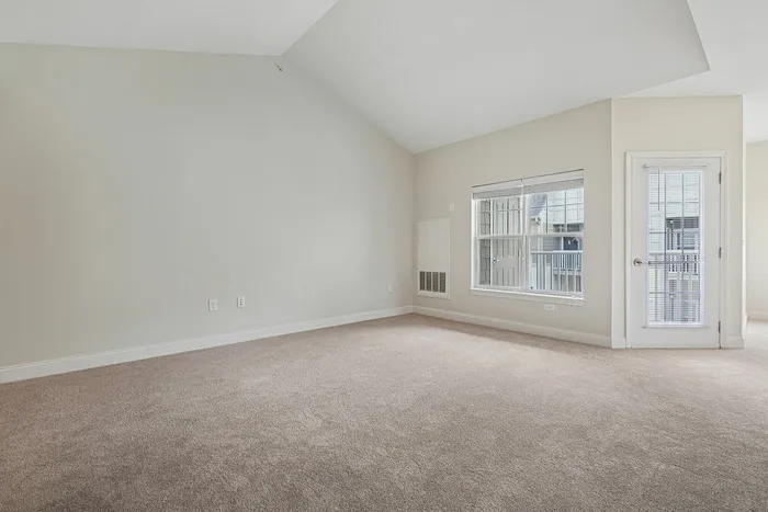 A spacious, empty apartment featuring a vaulted ceiling, beige carpet, large windows, and a glass door leading outside at our welcoming apartments in Westford, MA. | Princeton Westford