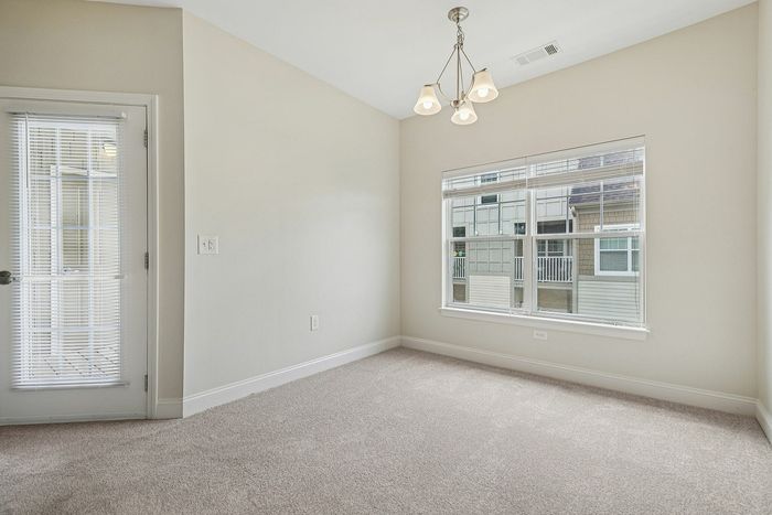 A bright, empty apartment in Westford, MA with beige walls and carpet, a window overlooking nearby buildings, a glass door with blinds, a modern ceiling light fixture, and space perfect for a cozy breakfast nook. | Apartments with Balcony in Westford