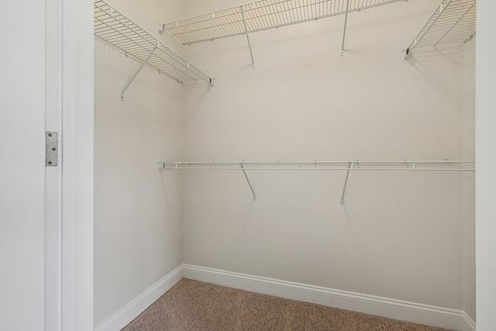 Empty walk-in closet with beige carpet, white walls, and multiple white wire shelves makes an ideal linen closet in this apartment in Westford, MA. | Apartments with Balcony in Westford