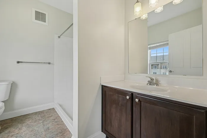 Modern bathroom in an apartment in Westford, MA, featuring a dark wood vanity, white countertop, large mirror, sink, and walk-in shower. Light-colored walls complement the space with a towel bar and a partially visible toilet on the left. | Apartments with Air Conditioning in Westford