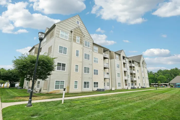 A light-colored, four-story Princeton Westford Apartment Community in Westford, MA, features balconies and gabled roofs, surrounded by green lawns, trees, and a sidewalk under a blue sky with scattered clouds. | Pet Friendly Apartments in Westford