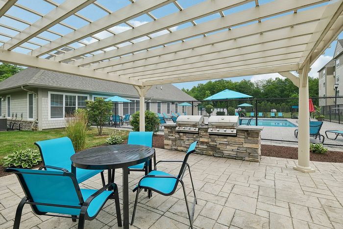 Outdoor patio area featuring seating under a pergola, a community grilling area, and a fenced swimming pool nearby at our welcoming apartments in Westford, MA. | Princeton Westford Apartments | Wheelchair Accessible Apartments in Westford