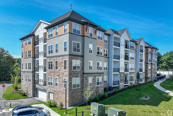 The James Apartments in Haverhill MA is a modern, four-story building with stone and siding exterior, large windows, and balconies, nestled among green grass, trees, and a clear blue sky. | The James | Apartment in Haverhill, MA