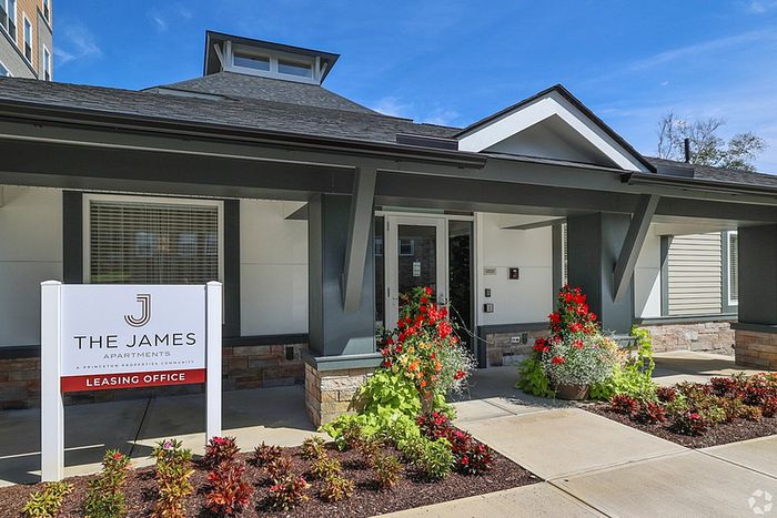 Modern apartment leasing office exterior in Haverhill MA, featuring gray siding, large windows, and a peaked roof. A sign out front reads The James Apartments Leasing Office, with colorful flowers and neatly trimmed bushes lining the walkway. |The James | Apartment in Haverhill, MA