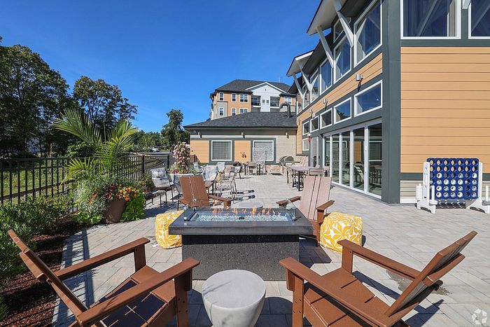 Outdoor patio at The James Apartments in Haverhill, MA features lounge chairs around a modern fire pit, surrounded by tables, potted plants, and a giant Connect Four game beside a tan and green building under a clear blue sky. | The James | Apartment in Haverhill, MA