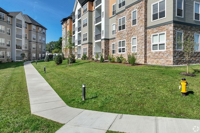 The James Apartments in Haverhill MA features a modern beige and stone exterior, green lawns, small shrubs, a yellow fire hydrant, and a curved sidewalk under a clear blue sky. | The James | Apartment in Haverhill, MA