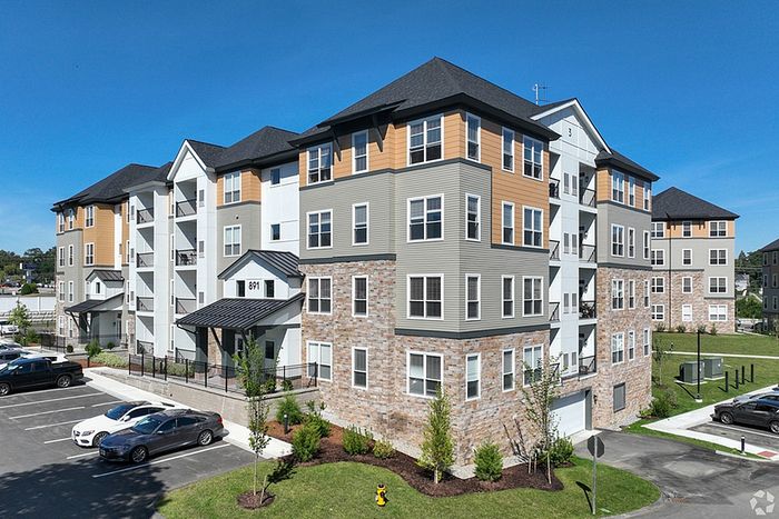 The James Apartments in Haverhill MA is a modern four-story building with gray and tan siding, large windows, and stone accents, surrounded by landscaped areas and parked cars under a clear blue sky. | The James | Apartment in Haverhill, MA