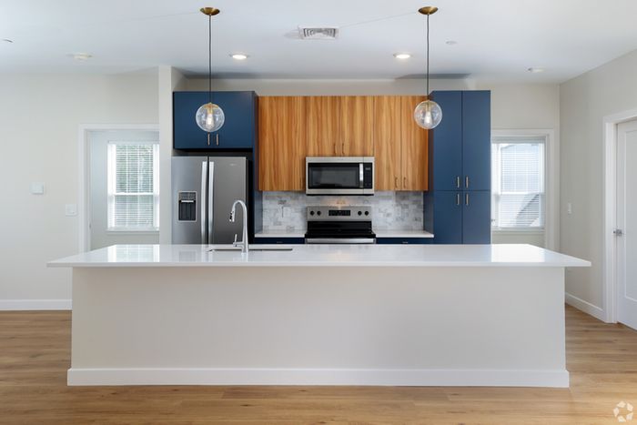 Modern kitchen at The James Apartments in Haverhill MA, featuring a large white island, stainless steel appliances, wood and blue cabinetry, globe pendant lights, light wood flooring, and windows on either side. | The James | Apartment in, Haverhill MA