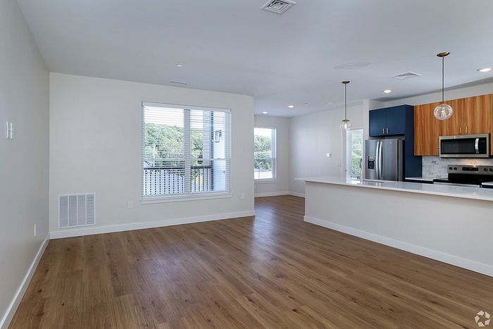 Modern, unfurnished open-plan living space at The James Apartments in Haverhill MA, featuring light wood flooring, large windows, and a kitchen with blue and wood cabinetry, stainless steel appliances, and white countertops.  | The James | Apartment in, Haverhill MA
