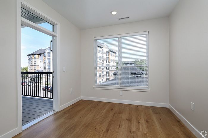 A bright, empty room at The James Apartments with light wood flooring, white walls, a large window with blinds, and a glass door to a balcony overlooking an apartment building in Haverhill MA. | Apartments with Balcony in Haverhill | The James | Apartment in, Haverhill MA