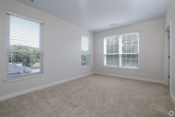 A bright, empty room at The James Apartments in Haverhill, MA features beige carpet, white walls, and three large windows with white blinds letting in natural light. A small doorway is to the right.  | The James | Apartment in, Haverhill MA