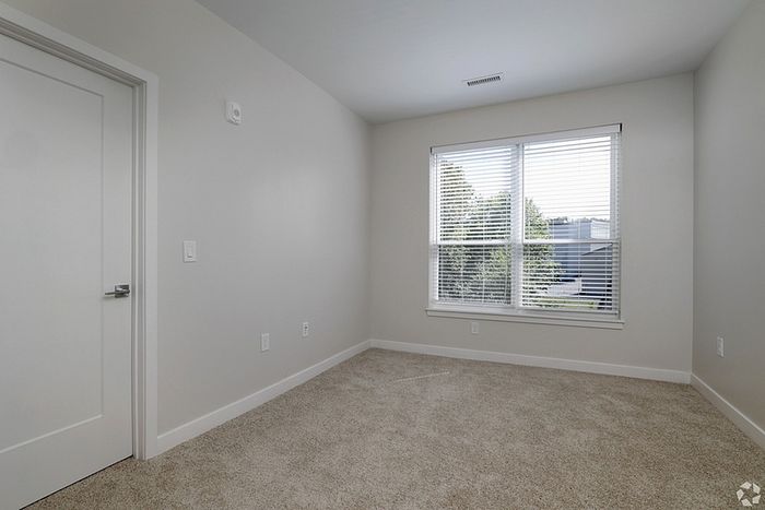 An empty room at The James Apartments in Haverhill, MA features beige carpet, light gray walls, a white door, and a large window with white blinds letting in natural light. Trees and neighboring buildings are visible outside the window.  | The James | Apartment in, Haverhill MA