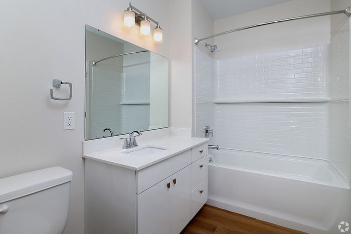 Modern bathroom at The James Apartments in Haverhill, MA, featuring a white vanity, rectangular mirror, three-light fixture, towel ring, toilet, and a bathtub with white subway tile and curved shower curtain rod. Wood-style flooring adds warmth.  | The James | Apartment in, Haverhill MA