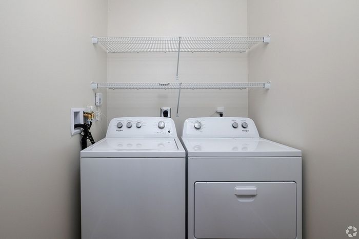 A laundry room at The James Apartments in Haverhill MA features a white washing machine and dryer side by side against a beige wall, below two empty white wire shelves. | The James | Apartment in, Haverhill MA