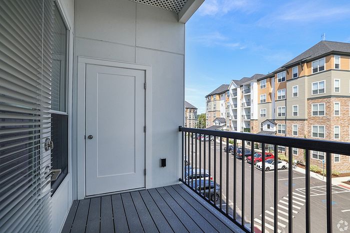 A modern apartment balcony at The James Apartments in Haverhill, MA, features gray flooring, a white door, metal railing, and a view of the parking lot and multi-story buildings on a sunny day  | The James | Apartment in, Haverhill MA