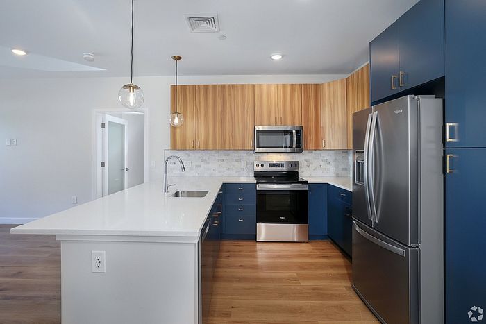 Modern kitchen at The James Apartments in Haverhill MA features stainless steel appliances, blue lower cabinets, wooden uppers, white quartz countertops, tile backsplash, pendant lights over a peninsula island, and light wood flooring throughout.  | The James | Apartment in, Haverhill MA