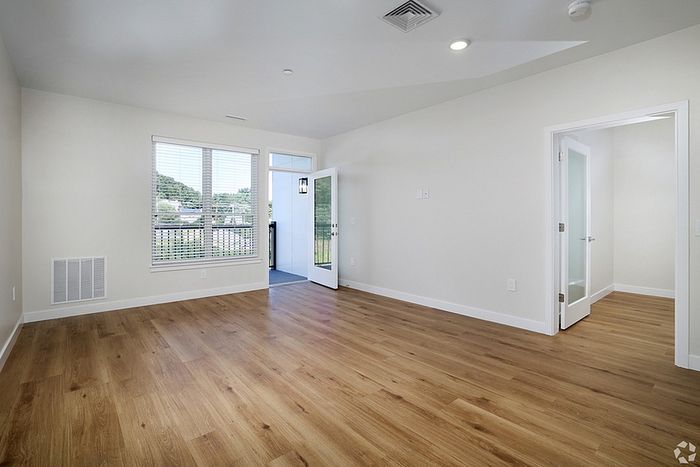 A bright, empty room at The James Apartments in Haverhill MA features light wood flooring, white walls, a large window, a glass door to the balcony, and another door with frosted glass panels leading to a separate space.  | The James | Apartment in, Haverhill MA