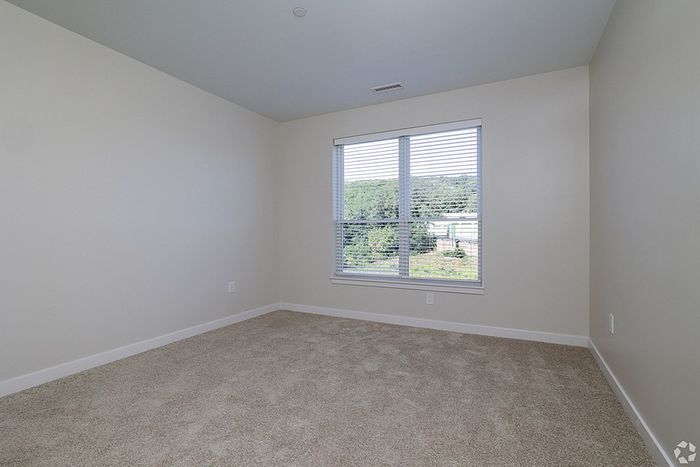 Empty room at The James Apartments in Haverhill, MA featuring beige carpet, light walls, and a large window with white blinds allowing natural light in. Enjoy views of trees and greenery just outside your window.  | The James | Apartment in, Haverhill MA