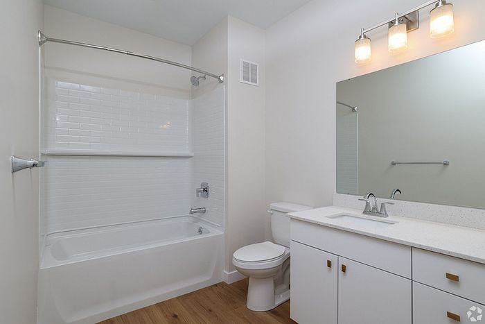 Modern bathroom at The James Apartments in Haverhill MA featuring white walls, a bathtub with white subway tiles, a toilet, sleek vanity with sink, large mirror, and three-light fixture above. Wood-like flooring completes the contemporary design.  | The James | Apartment in, Haverhill MA