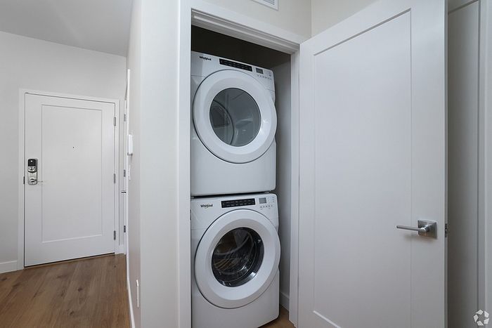 A stacked white washer and dryer set is installed in a small laundry closet with white walls and a partially open door, located in the hallway of The James Apartments in Haverhill MA, featuring modern wood flooring.  | The James | Apartment in, Haverhill MA
