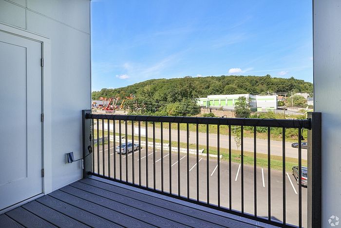 A view from a balcony with black railings at The James Apartments in Haverhill, MA, overlooking a parking lot, road, and green buildings set against a tree-covered hill under a clear blue sky.  | The James | Apartment in, Haverhill MA