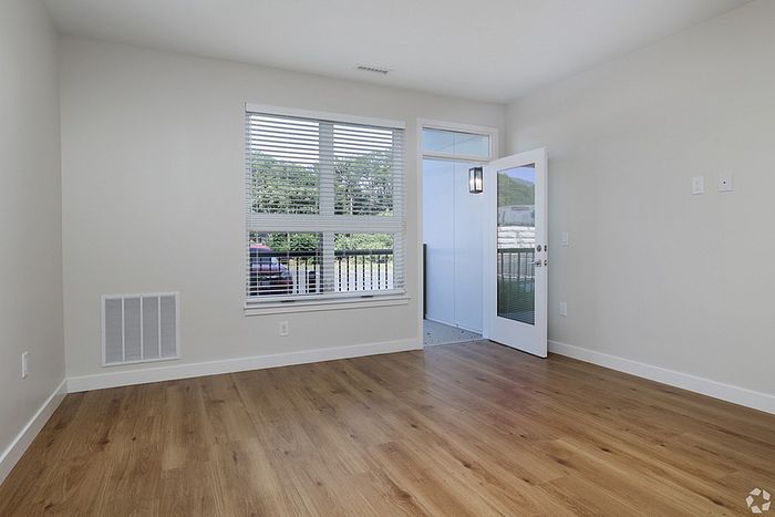 Bright, empty room at The James Apartments in Haverhill MA with light-colored walls, wood flooring, a large window with blinds, and an open door leading to a balcony with black railing and green outdoor view.  | The James | Apartment in, Haverhill MA
