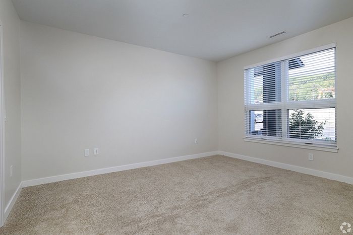 A bright, empty room at The James Apartments in Haverhill MA features beige carpet, white walls, a large window with blinds, and electrical outlets. Natural light streams in, revealing lush greenery outside.  | The James | Apartment in, Haverhill MA