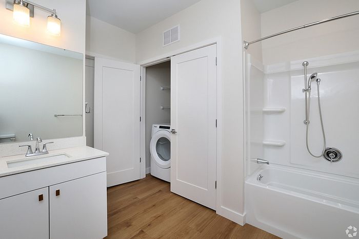 Modern bathroom at The James Apartments in Haverhill MA, featuring a white vanity, large mirror, bathtub with shower, and wood flooring. An open door reveals a washer and dryer in a convenient closet.  | The James | Apartment in, Haverhill MA