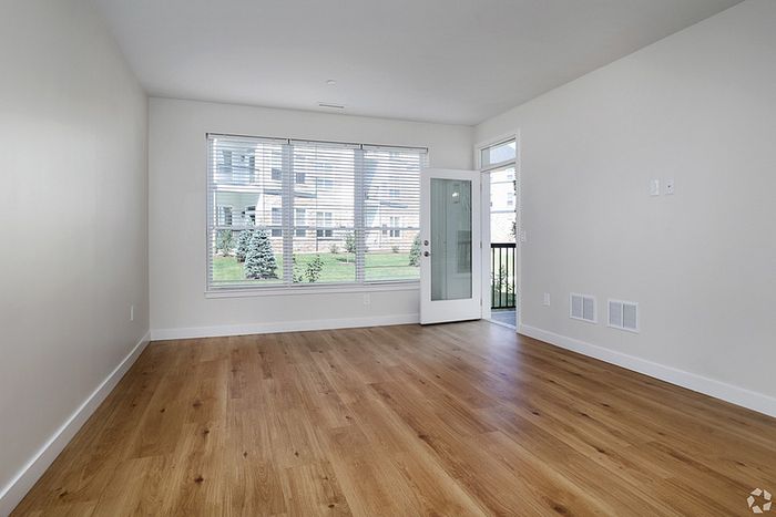 A bright, empty room at The James Apartments in Haverhill MA features light-colored walls, large windows with blinds, a glass door to the patio, and wood flooring. Natural light fills the space with views of a grassy outdoor area.  | The James | Apartment in, Haverhill MA