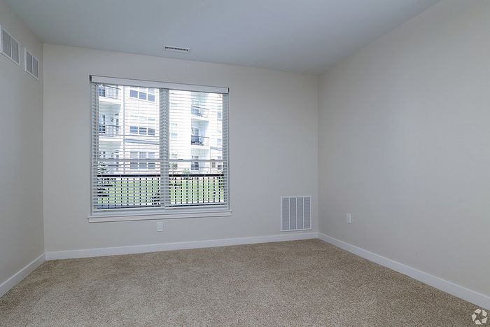 Empty room at The James Apartments in Haverhill, MA, featuring beige carpet, white baseboards, light walls, and a large window with blinds that fill the space with natural light. Apartment buildings are visible outside the window.  | The James | Apartment in, Haverhill MA