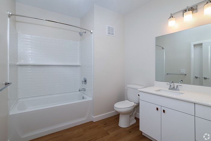 A modern bathroom at The James Apartments in Haverhill, MA, features a white bathtub and shower, a toilet, a sleek vanity with sink and mirror, three lights above the mirror, wood-style flooring, and light beige walls.  | The James | Apartment in, Haverhill MA