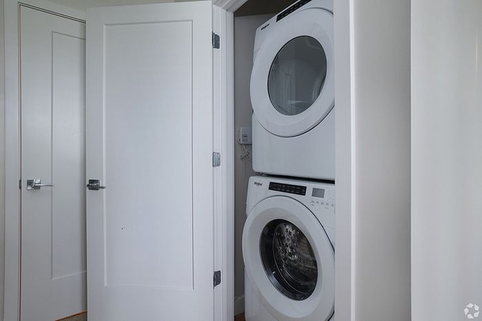 A stacked white washer and dryer set is installed in a small laundry nook at The James Apartments, Haverhill MA, with white walls and an open door. Another closed door is visible to the left.  | The James | Apartment in, Haverhill MA