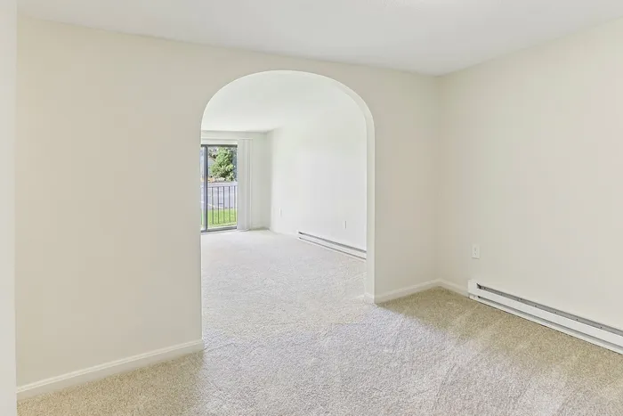 Princeton Place Apartments | A bright, empty room at Princeton Place Apartments in Worcester MA features light beige carpet, white walls, and an arched doorway. A sliding glass door in the background opens to a patio and grassy area outside. | Apartments with Washer & Dryer in Worcester