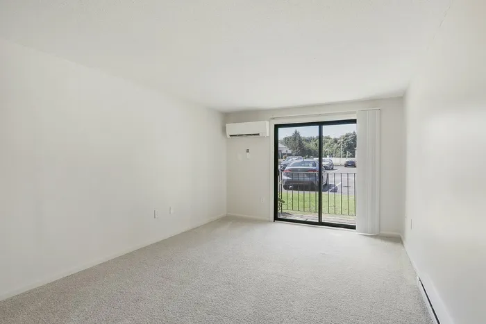 Princeton Place Apartments | A bright, empty room with light carpet and white walls at Princeton Place Apartments in Worcester MA, featuring a sliding glass door that opens to a patio with views of a grassy area and the parking lot. An air conditioning unit is mounted on the wall near the door. | Apartments with Balcony in Worcester