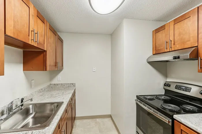 Princeton Place Apartments | A modern galley kitchen at Princeton Place Apartments in Worcester, MA, features wooden cabinets, light marbled countertops, a stainless-steel sink, four-burner electric stove, oven, vent hood, all under a bright ceiling light. | Worcester Apartments with Pickleball Court