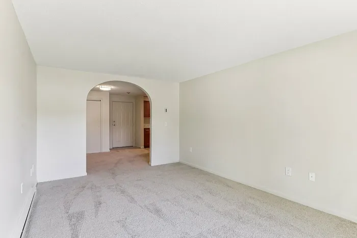 Princeton Place Apartments | Empty living room with light beige carpet and off-white walls at Princeton Place Apartments, featuring an arched doorway leading to a hallway and kitchen area in the background. The space appears clean and well-lit in Worcester MA. | Apartments with Washer & Dryer in Worcester