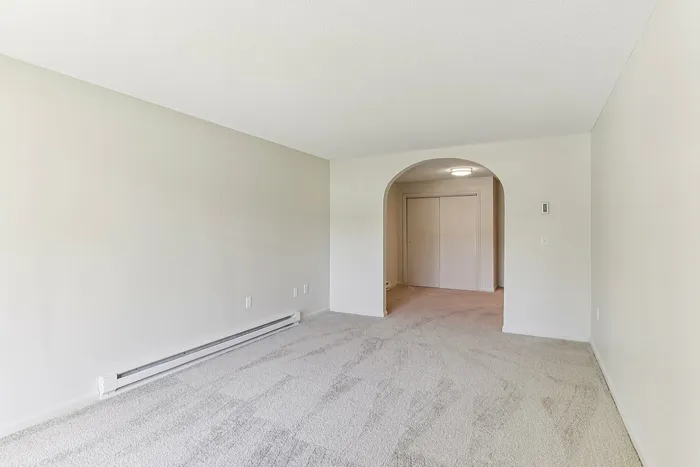 Princeton Place Apartments | Empty room at Princeton Place Apartments in Worcester MA, featuring light-colored carpet, off-white walls, a large archway, baseboard heater, and a closed closet with double doors under a ceiling light. | Wheelchair Accessible Apartments in Worcester