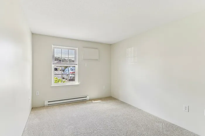 Princeton Place Apartments | A small, empty room with beige carpet, white walls, and a window letting in natural light at Princeton Place Apartments in Worcester MA. Enjoy a baseboard heater under the window and views of parked cars and greenery outside. | Apartments with EV Charging in Worcester