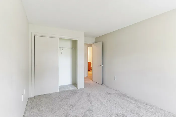 Princeton Place Apartments | Empty bedroom with light beige walls, an open white door, a closet with sliding doors, and light gray carpet flooring at Princeton Place Apartments in Worcester, MA. The room is well lit with natural light. | Apartments with Balcony in Worcester