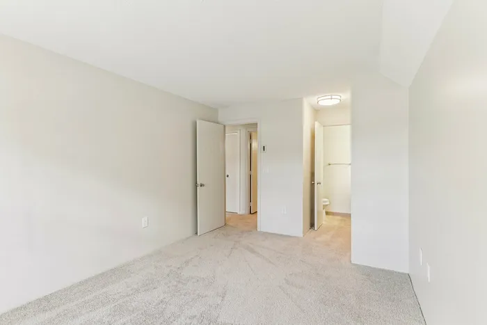 Princeton Place Apartments | A bright, empty room at Princeton Place Apartments in Worcester MA features beige carpet, white walls, an open door, and a view into a small bathroom with a toilet, closet space, and visible ceiling light fixture. | Wheelchair Accessible Apartments in Worcester