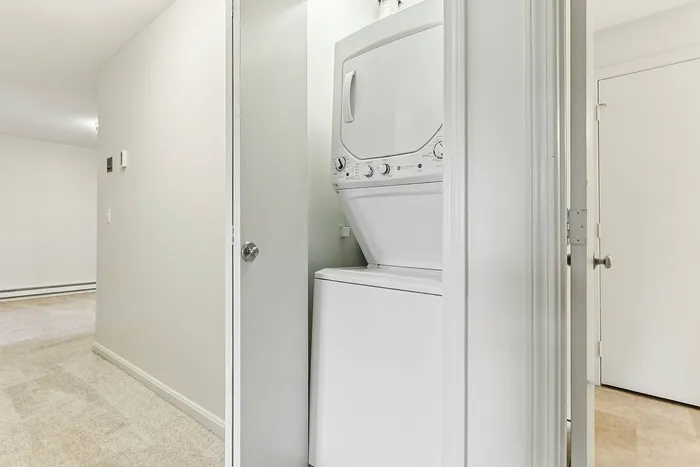 Princeton Place Apartments | A stacked white washer and dryer set is neatly placed inside a small laundry closet with an open door in the carpeted hallway at Princeton Place Apartments in Worcester MA. | Apartments with Washer & Dryer in Worcester