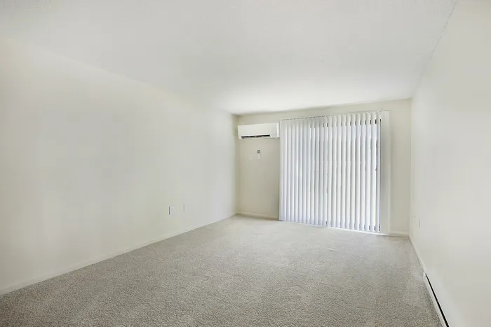 Princeton Place Apartments | Spacious empty room at Princeton Place Apartments in Worcester MA features white walls, beige carpet, a wall-mounted air conditioning unit, and large sliding glass doors with vertical blinds that let in natural light. | Apartments with Balcony in Worcester