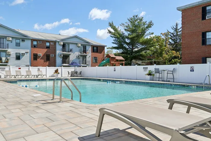 Princeton Place Apartments | A clean outdoor swimming pool at Princeton Place Apartments in Worcester MA is surrounded by a paved deck with lounge chairs, a white privacy fence, and apartment buildings in the background under a partly cloudy sky. | Apartments with Balcony in Worcester