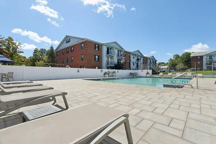 Princeton Place Apartments | Outdoor swimming pool area with lounge chairs, surrounded by a white fence, in front of the three-story Princeton Place Apartments in Worcester, MA, on a sunny day with a clear blue sky. | Apartments with Air Conditioning in Worcester