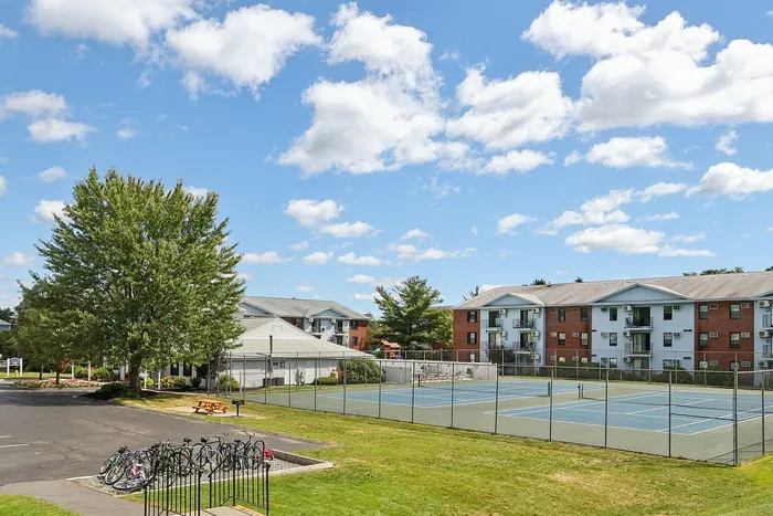 Princeton Place Apartments | A sunny outdoor scene at Princeton Place Apartments in Worcester MA, featuring tennis courts surrounded by a chain-link fence, apartment buildings in the background, a few trees, a picnic table, and a bike rack with several bicycles nearby. | Worcester Apartments with Pickleball Court