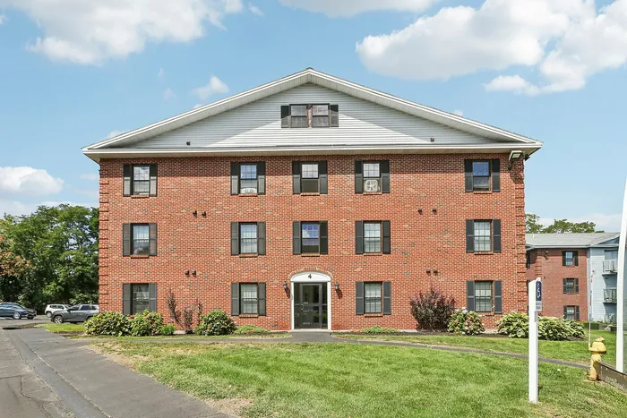 Princeton Place Apartments | Princeton Place Apartments is a three-story red brick building with white trim and a peaked roof, featuring several windows and surrounded by green lawns and shrubs in Worcester MA on a sunny day with blue sky and clouds. | Worcester Apartments with Virtual Tours